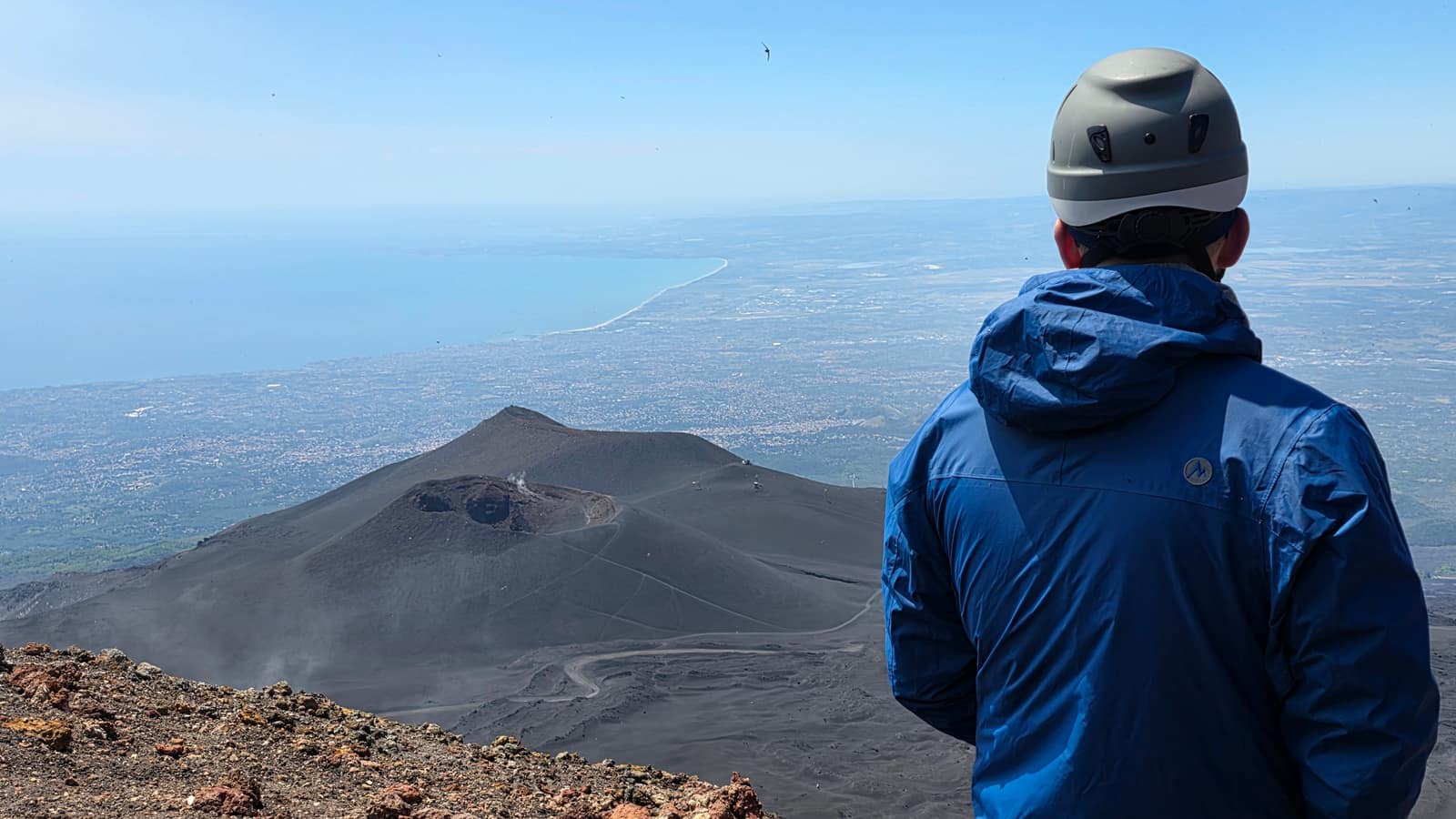 Ein Besucher mit Helm blickt über die Krater des Ätna auf Catania und die Küste