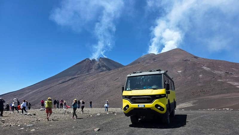 Tourists exploring Mount Etna’s upper slopes near active summit craters, with a yellow 4x4 off-road bus parked on volcanic terrain and volcanic smoke rising in the background on a Etna volcano tours